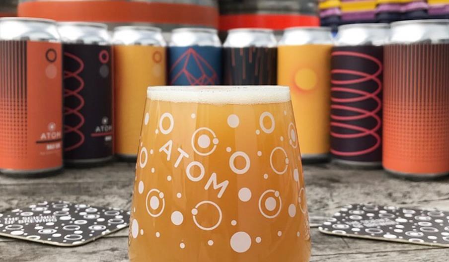 A glass of beer with the Atom logo on surrounded by colouful beer cans.
