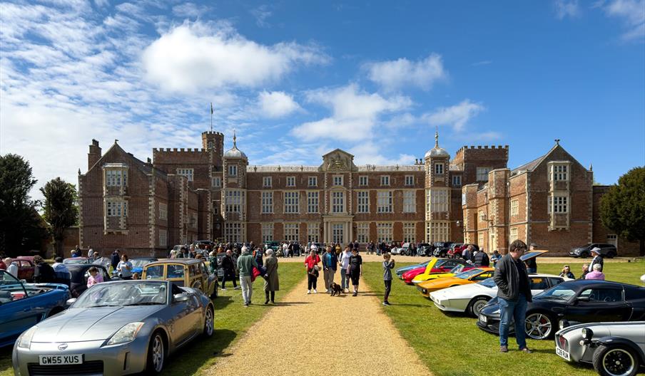 People are walking along a pathway lined with parked cars on a lawn in front of a large, ornate historic building under a partly cloudy sky.