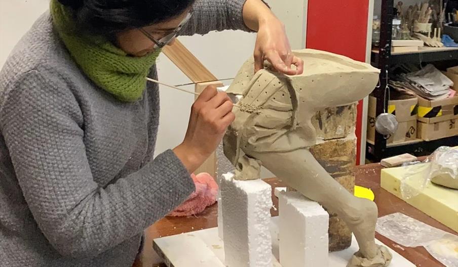 A person with dark hair and glasses is sculpting a clay figure on a table, using a wooden tool to add detail. The sculpture appears to be of a person
