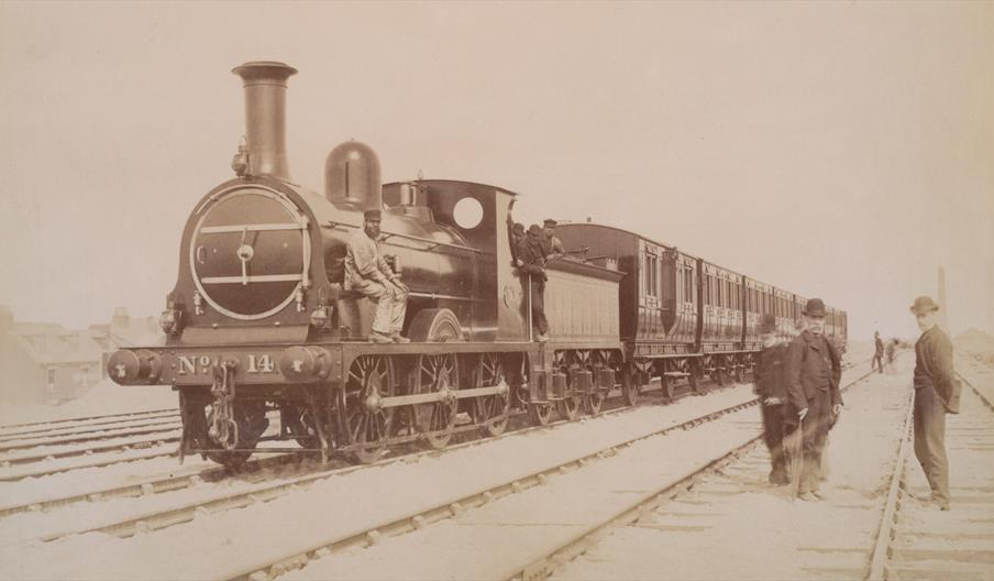 A sepia-toned photograph of a steam locomotive numbered 14 with several men posed on and beside the train, standing on railway tracks; passenger cars