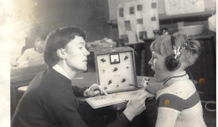A woman holds a microphone in front of a young boy wearing headphones, conducting a hearing or speech test with an old-fashioned audio testing device