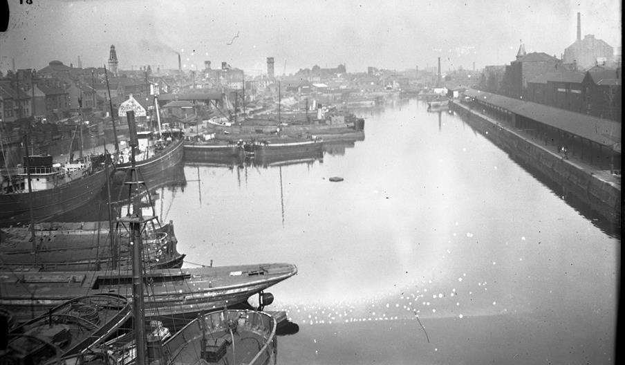 Black and white photo of an industrial canal lined with large docked ships on the left and a long warehouse on the right, with factories and smokestac