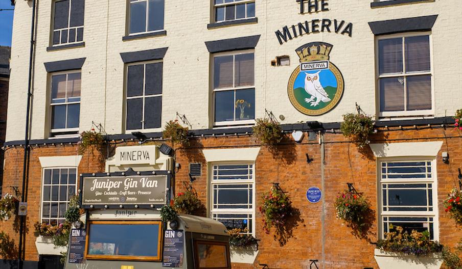 A vintage-style gin van is parked in front of The Minerva pub, a two-story brick building with flower boxes under the windows and a painted crest on t