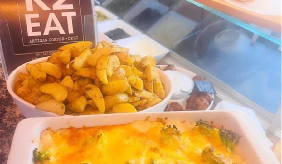 A close-up of a counter display at K2 Eat, featuring a white dish of baked cheesy broccoli casserole, a bowl of seasoned potato wedges, and a sign tha