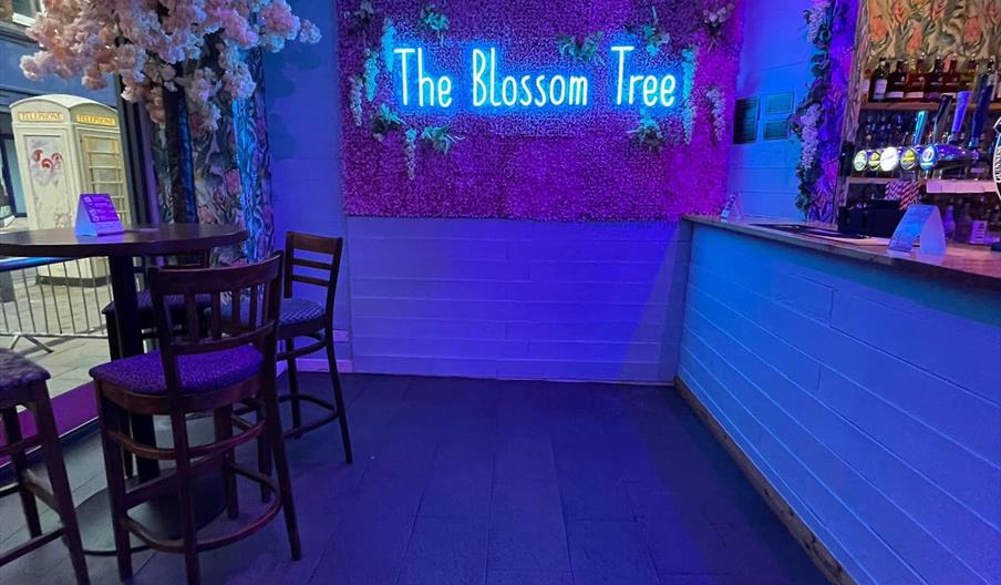 A bar area with purple and blue lighting features a neon sign reading "The Blossom Tree" mounted on a wall decorated with artificial flowers. To the l