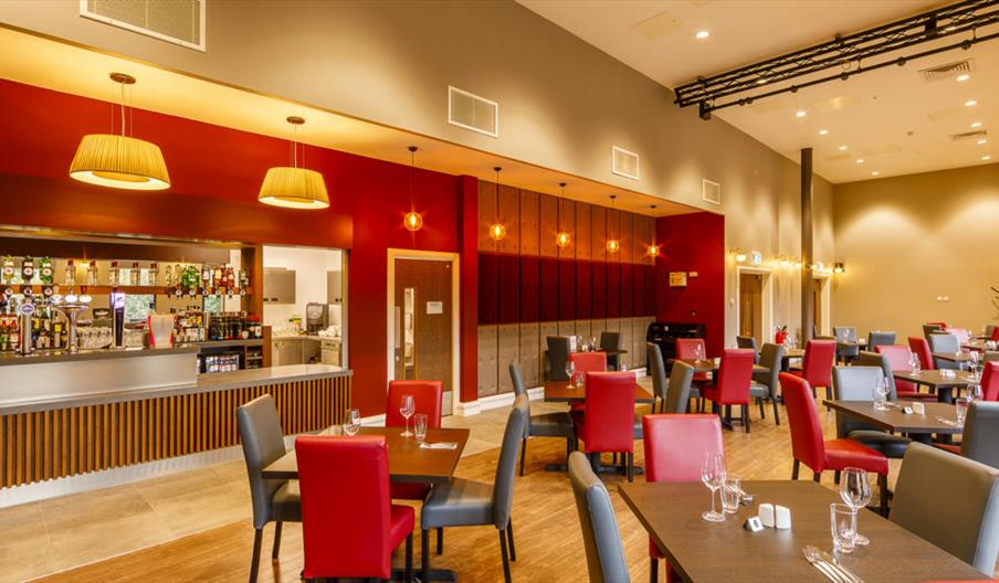 Interior of Hull New Theatre Lounge. A spacious, modern restaurant with a bar on the left, set against a red accent wall and illuminated by hanging la