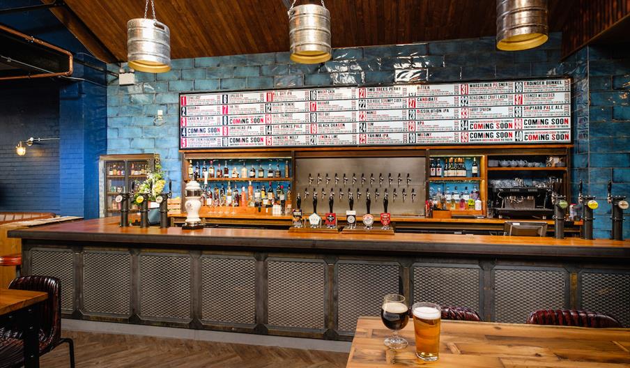Interior of Taphouse. A modern bar with a long counter, multiple beer taps, shelves of liquor bottles, and a large menu board listing various drinks o