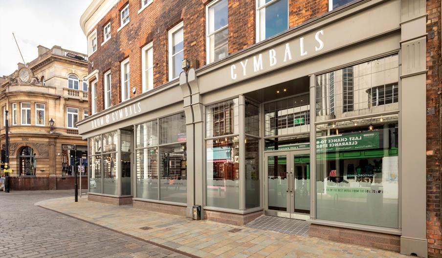 Street view of a corner building with large windows and a sign reading "DRUM & CYMBALS" above the entrance, located on a paved city street with histor