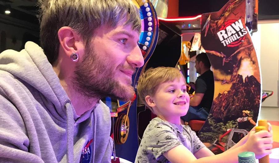 A man and child play an arcade game with arcade games in background.
