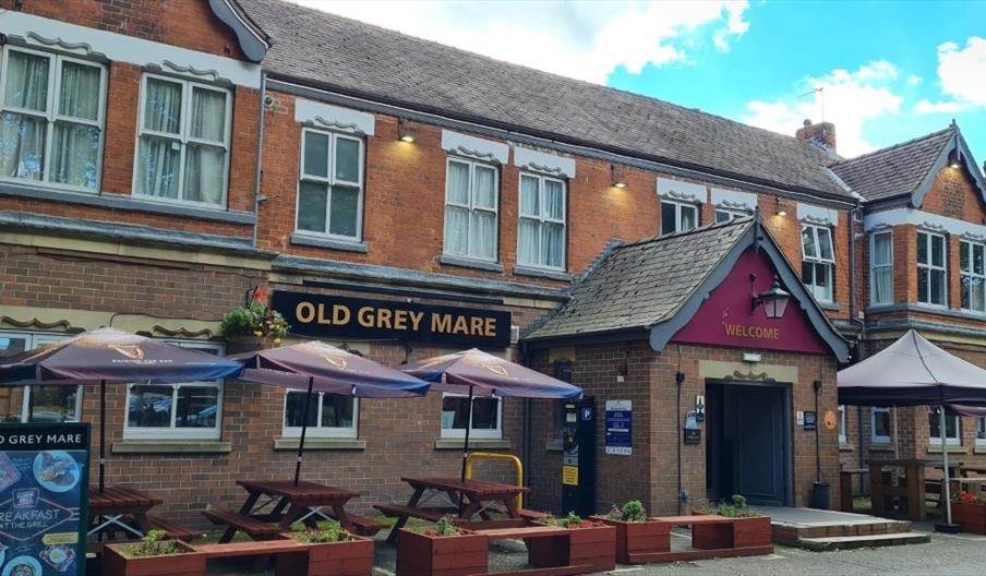 The exterior of the Old Grey Mare, a traditional brick pub with large windows and a sign above the entrance. There are several wooden picnic tables wi
