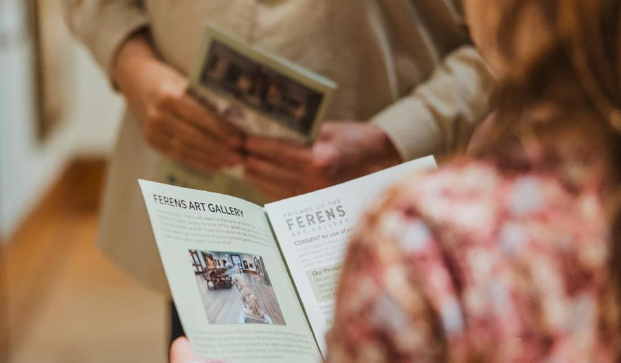 An over-the-shoulder shot of a person reading a programme on the contents of Ferens Art Gallery whilst another person stands in the background holding