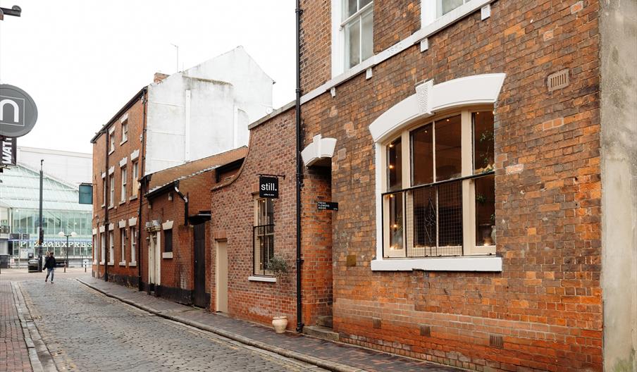 A narrow cobblestone street is lined with old brick buildings, including a café with large windows and a "Still." sign. In the background, a modern gl