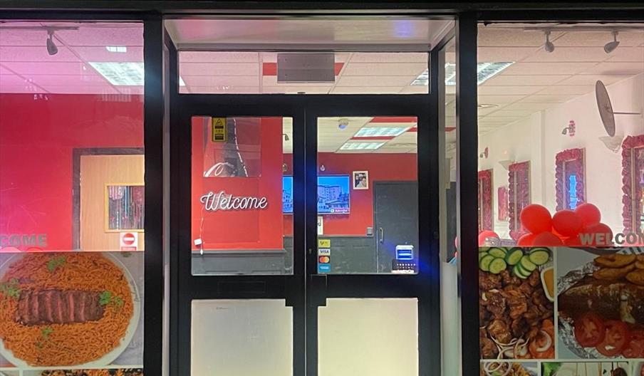 Front entrance of the brightly lit restaurant "Tantalizer Africana Adorable Kitchen," with "Welcome" on the door and photos of assorted African dishes