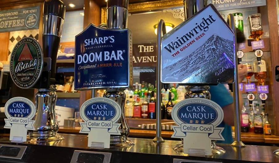 Interior of The Avenue. Three beer taps in a pub display labels for Bass, Sharp's Doom Bar, and Wainwright the Golden Beer, with "Cask Marque Cellar C