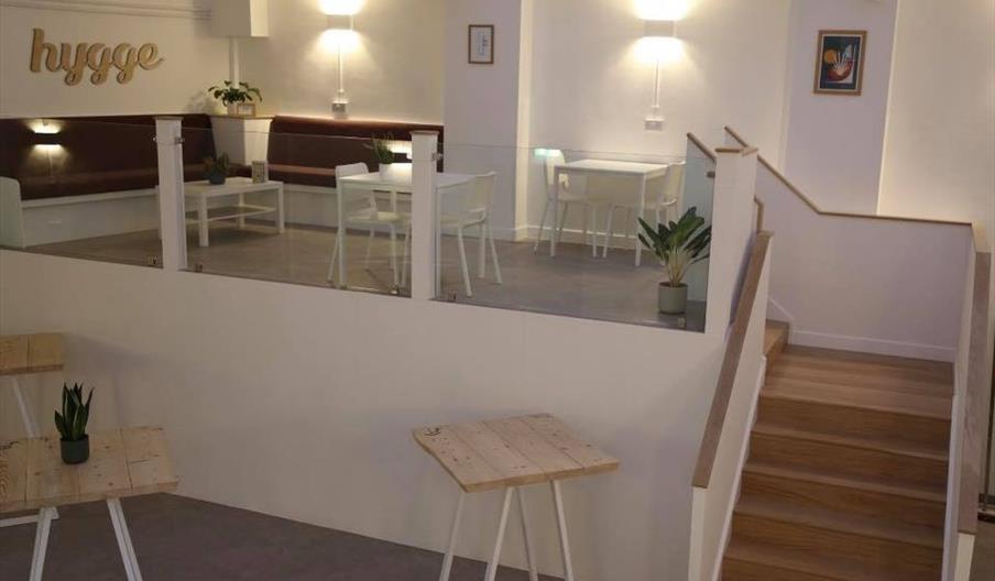 Interior of Hygge. A modern, minimalist café interior with light wooden tables, white chairs, potted plants, and a staircase leading up to a seating a