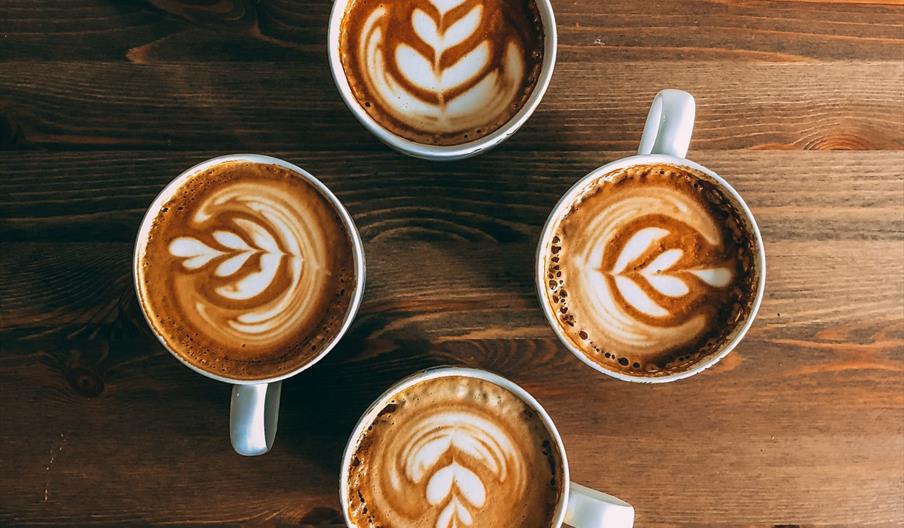 Four cups of latte with leaf-shaped latte art sit arranged in a circle on a wooden table, seen from above.