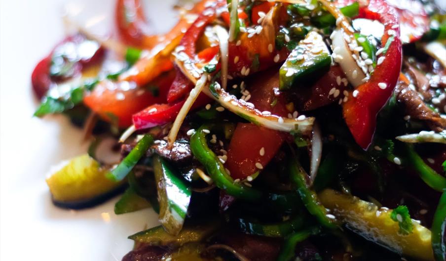 A colorful vegetable salad on a white plate, featuring sliced cucumbers, red bell peppers, tomatoes, and green herbs, garnished with sesame seeds. The
