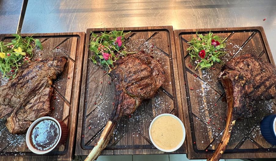 Three large grilled tomahawk steaks are served on wooden boards, each with a side of sauce and a garnish of greens and edible flowers. The steaks are