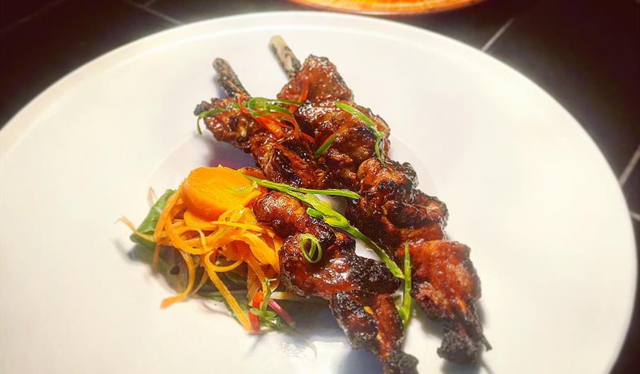 Three grilled meat skewers garnished with sliced green onions are served on a large white plate with a small side of shredded carrots and other vegeta