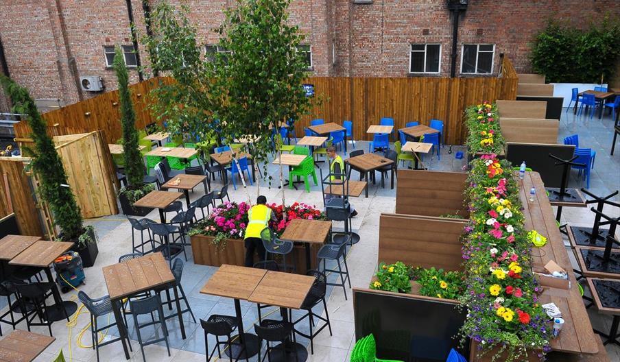 Outdoor seating area with several tables and chairs, some greenery, and a person tending to a planter with flowers. Brick walls and wooden fencing enc