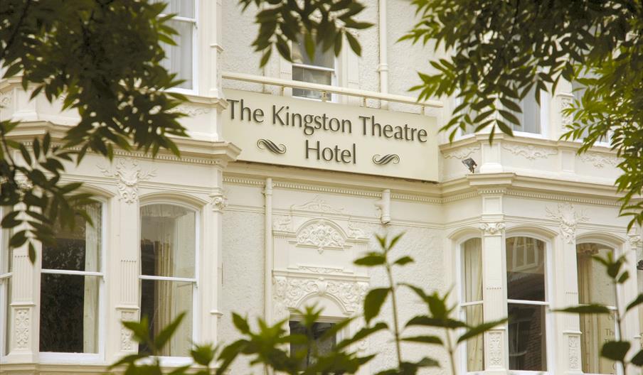 Exterior of white, ornate building flanked by trees with a gold sign reading Kingston Theatre Hotel.