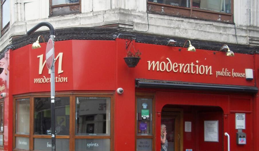 A corner view of a red-painted pub called "Moderation public house" with large windows, gold signage, and a person standing just inside the entrance.