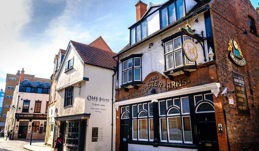 A cobblestone street lined with historic English buildings, including pubs and eateries with signs reading "The Manchester Arms," "Old House," and "Li