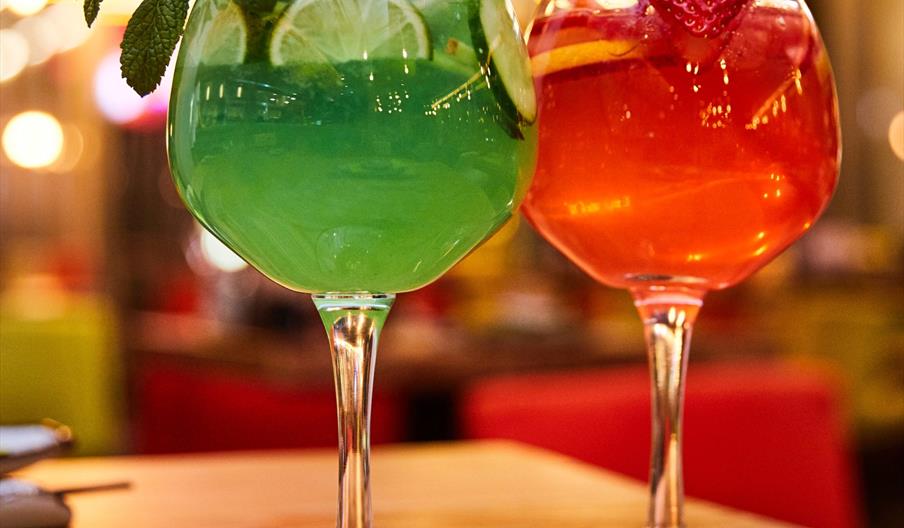 Two cocktails on a wooden bar one green with mint and one red with strawberrys both with straws.