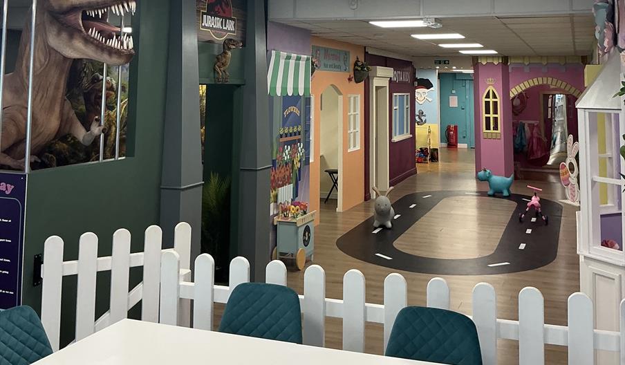 A seating area sits in front, with a fence separating it from a playarea featuring several fake shopfronts, a circular road playmat and several childr