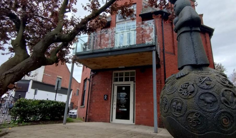 The front entrance of 87 Gallery, a redbrick building with a glass edged balcony. A large tree looms over an art installation outside.