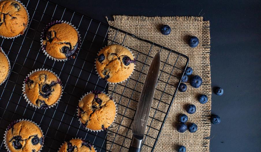 Several blueberry muffins are cooling on a wire rack, with a knife, fresh blueberries, and a piece of burlap on a dark tabletop.