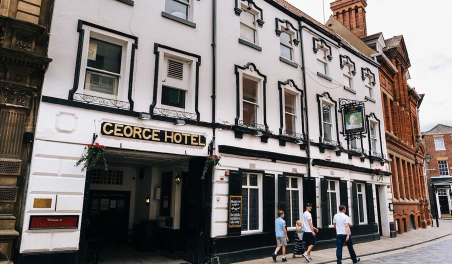 A white, historic-looking building with a sign reading "George Hotel" above an entrance; four people are walking on the cobblestone street in front of