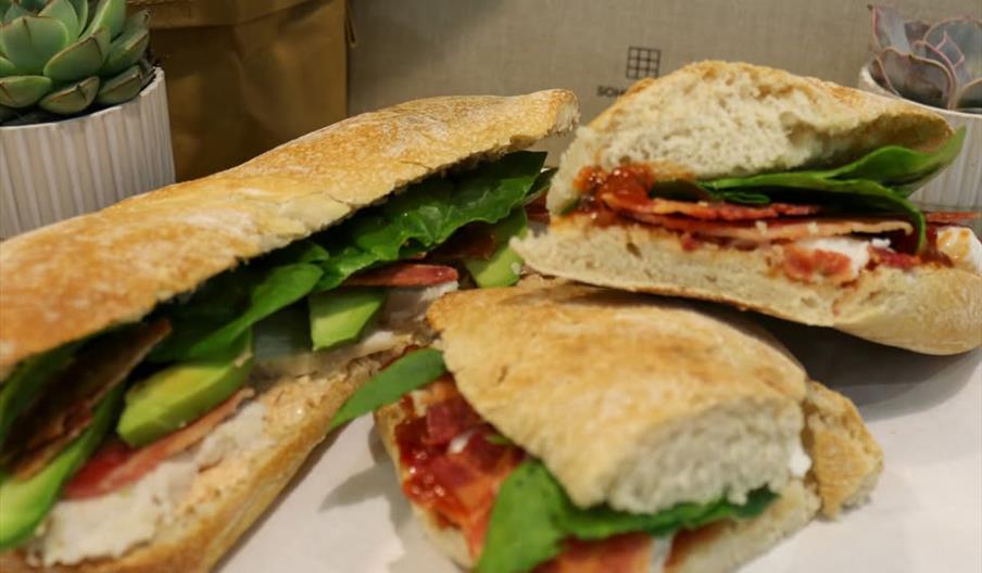 Three sandwiches on white paper, the largest being an open sandwich, filled with sliced avocado, lettuce, and bacon. Two smaller sandwich halves are p