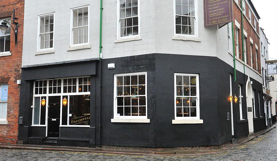 A corner building with black-painted lower walls and white upper walls, featuring large windows and glowing lanterns by the entrance; the building is