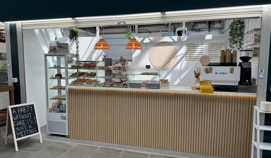 A stall in Trinity Market, with wooden panelled front, glass cabinets displaying various cakes, coffee machine on the counter and a person in the back