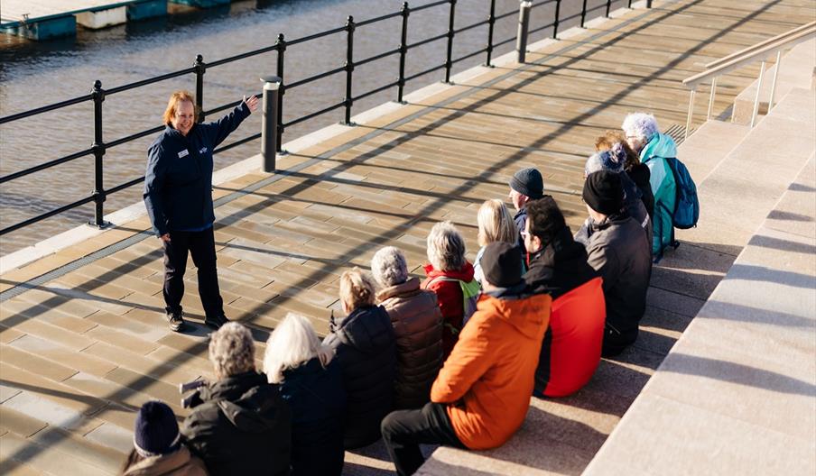 A tourguide gesticulates in front of a crowd of people sat on a series of steps facing the Hull Marina.