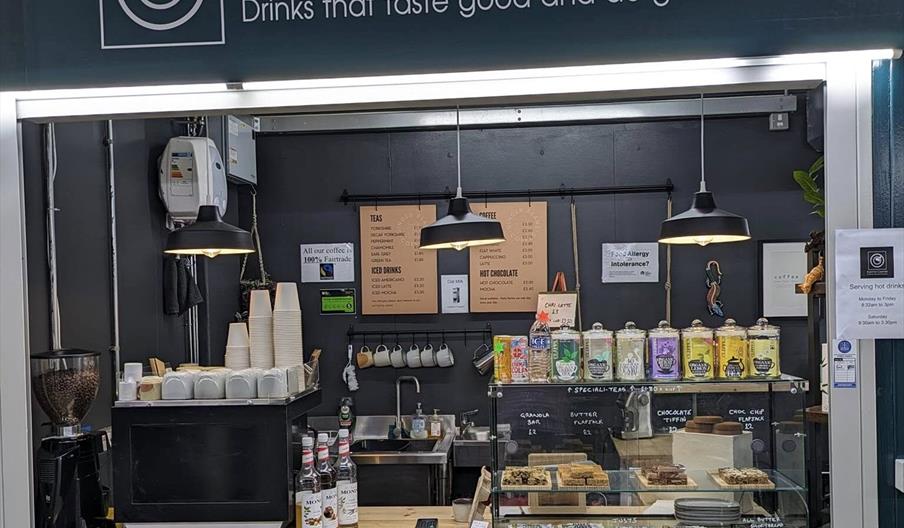 The small café kiosk of Karma Corner, with a sign reading "Drinks that taste good and do good," features a counter displaying baked goods, stacked cup