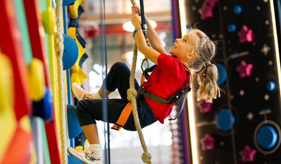 A small child in a harness smiles as they pull a rope to help themselves climb a multicoloured climbing wall.