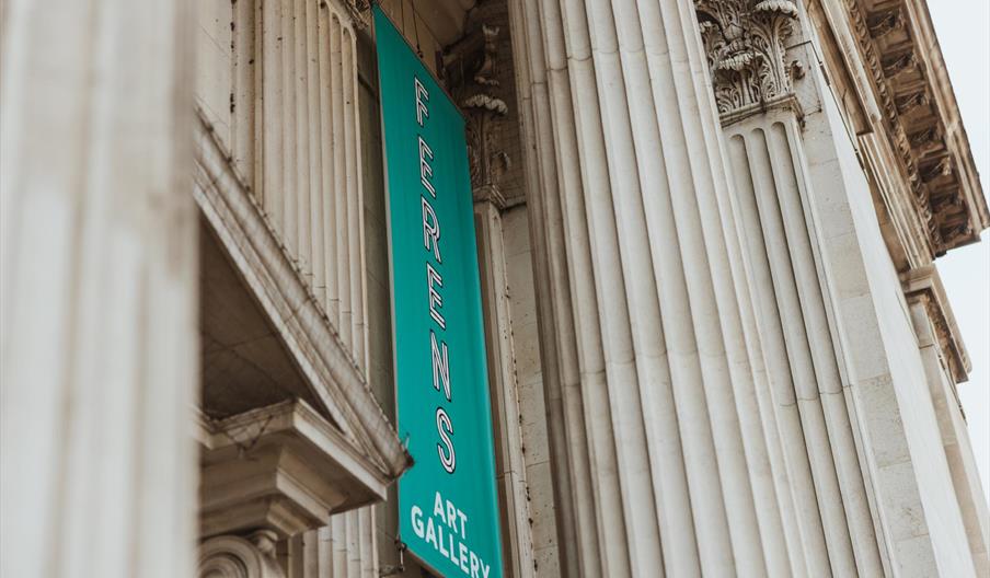 A tall turquoise banner reading "Ferens Art Gallery" hangs between large ornate stone columns on the facade of a grand neoclassical building.