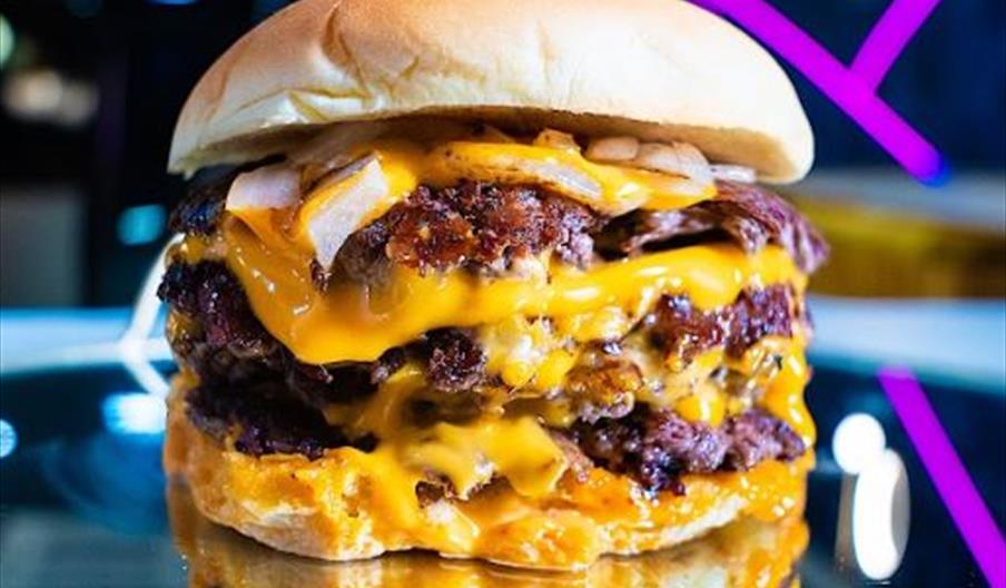A close-up of a large cheeseburger with multiple beef patties, melted cheese, and chopped onions, reflected on a glossy surface with colorful lights i