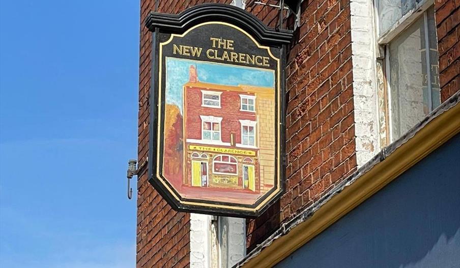 A hanging sign on a brick building reads "The New Clarence" and features a painted image of a red-brick pub façade under a blue sky.