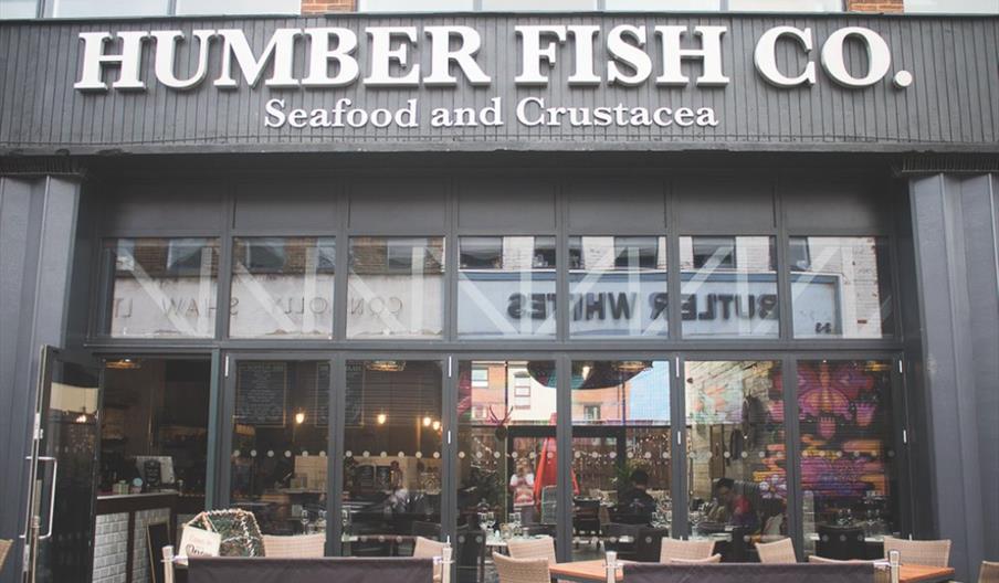 Exterior of Humber Fish Co., a seafood and crustacea restaurant. Large glass windows and doors reveal the dining area inside, with tables and chairs s