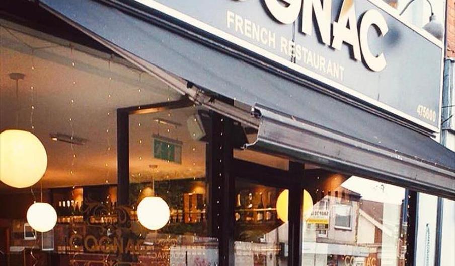 Exterior of restaurant with plant display in planters, canopy and sign in black and gold reading Cognac French Restaurant.