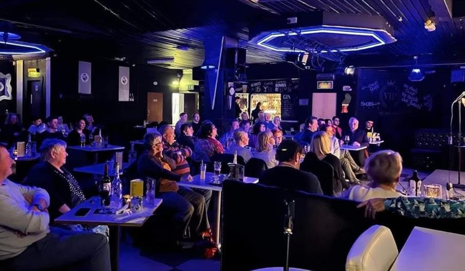 Crowd sat at tables inside a comedy club laughing while watching a performance on stage.