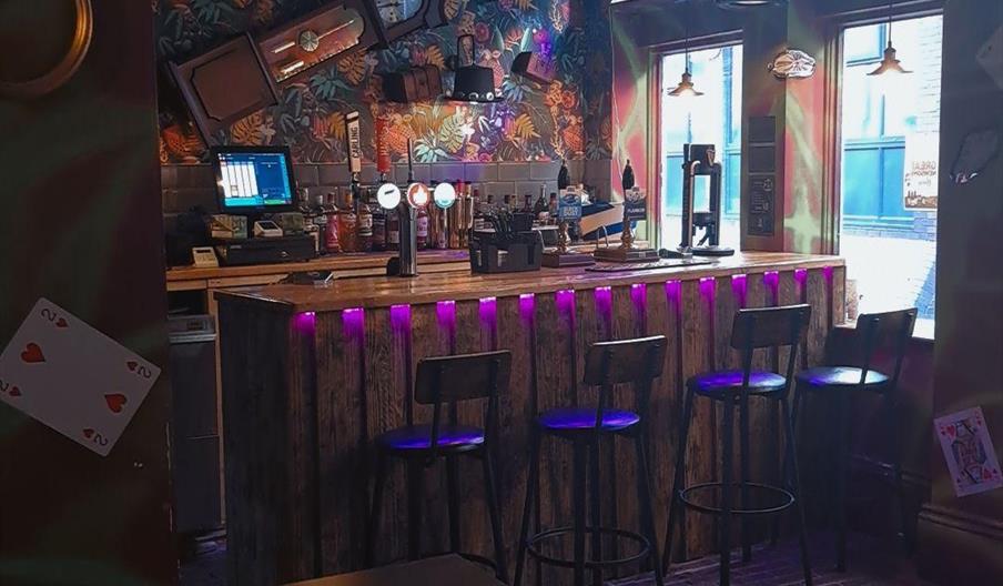 Interior of Hatters Cafe Bar. A whimsical bar with a wooden counter illuminated by purple lights, four high chairs, and a colorful, patterned wall fea