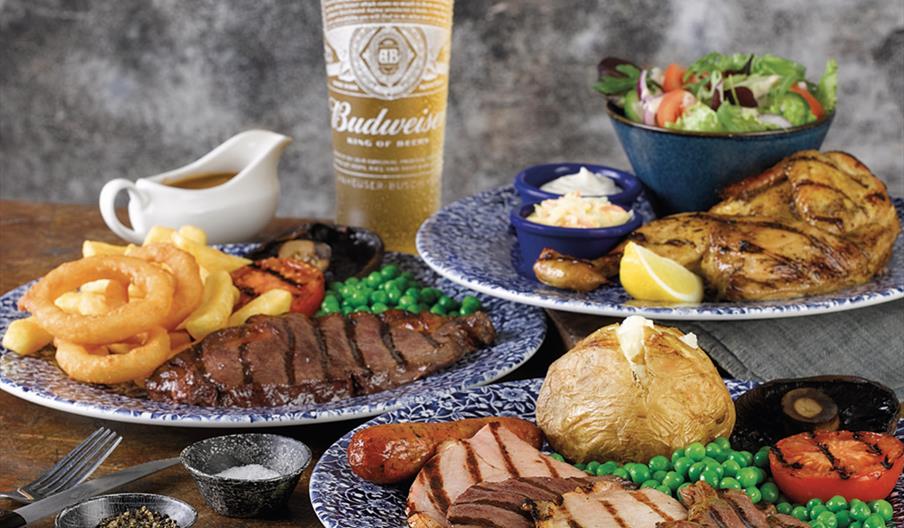 Three plates on a wooden table, featuring a mixed grill of different meats, peas, grilled tomatoes and mushrooms, some salt and pepper in small pots,
