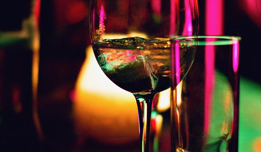 Bar of The Harry Pursey. A wine glass filled with ice and a clear liquid sits on a dimly lit bar, surrounded by colorful reflections and blurred glass