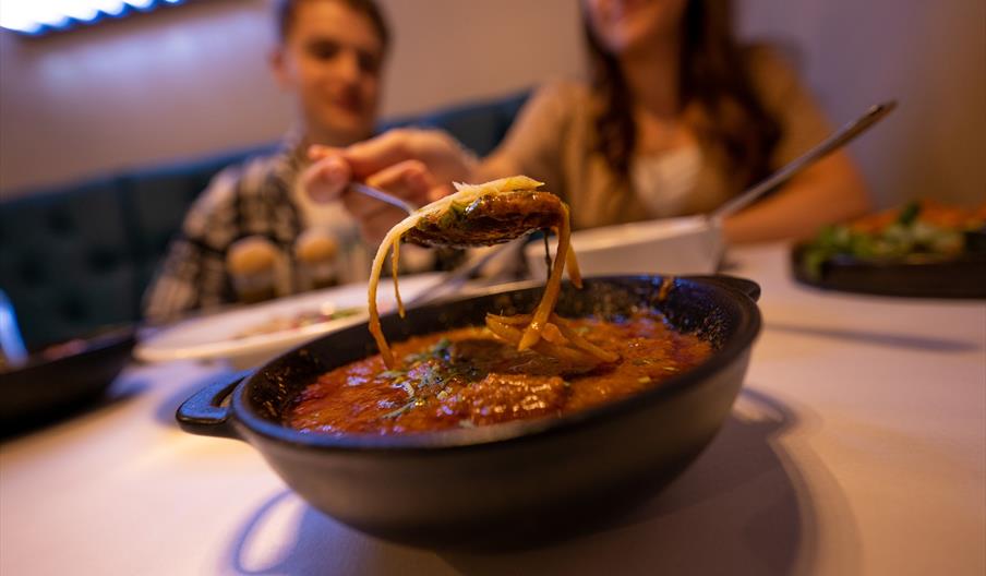 Curry in black bowl with spoon and couple in background.