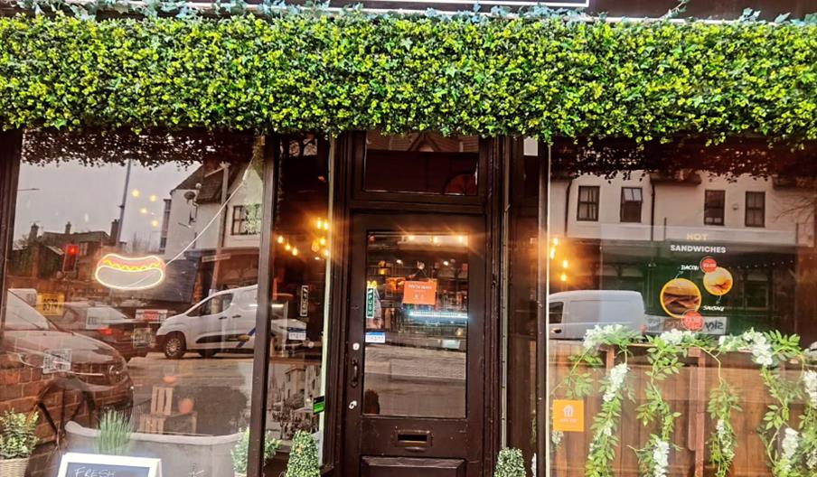 Storefront of the "Lunch Shop" with a black sign featuring a hot dog graphic, artificial green foliage above the entrance, two topiary plants by the d