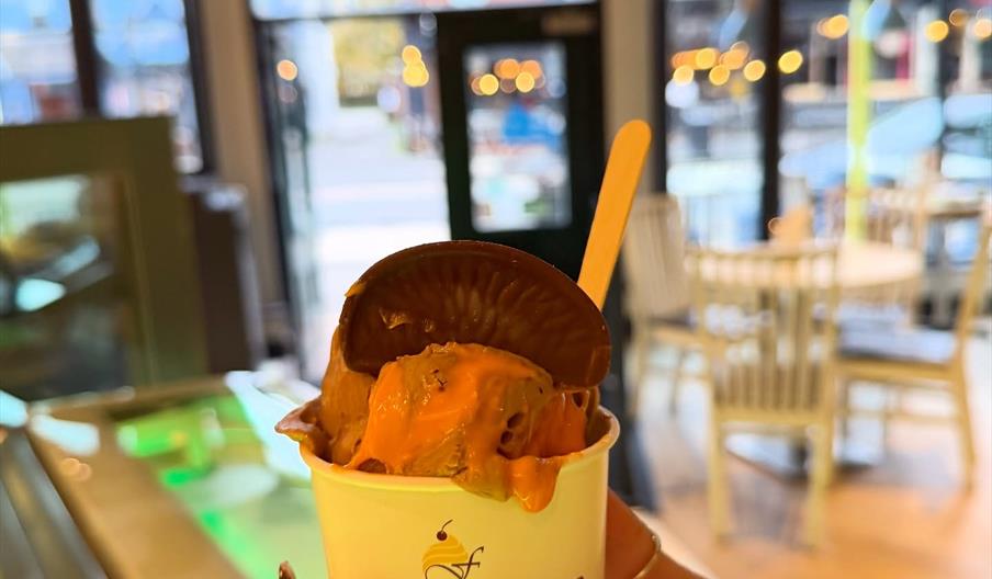 Chocolate ice cream in a paper tub with wooden spoon held in a persons hand.